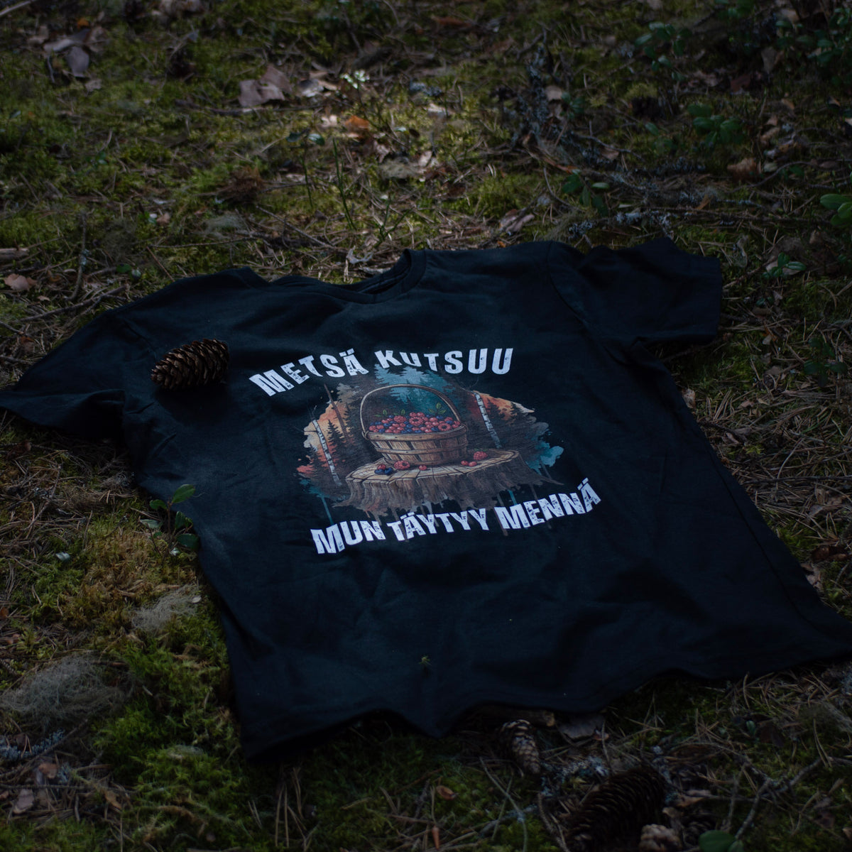 Musta Metsä kutsuu, mun täytyy mennä -t-paita marjakoriprintillä, aseteltuna metsämaahan.