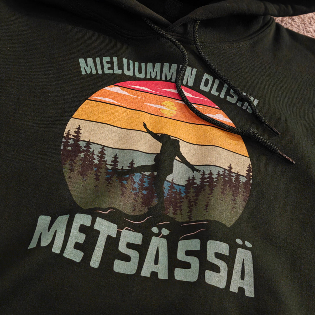Tumma Mieluummin olisin metsässä -huppari retkeilijän siluetilla ja auringonlaskuprintillä, lähikuvassa.