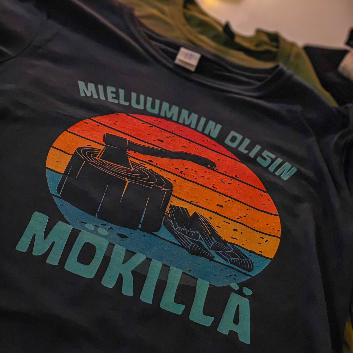 Tumma Mieluummin olisin mökillä -t-paita, jonka printissä on kirves ja klapeja, kuvattuna tasolla.