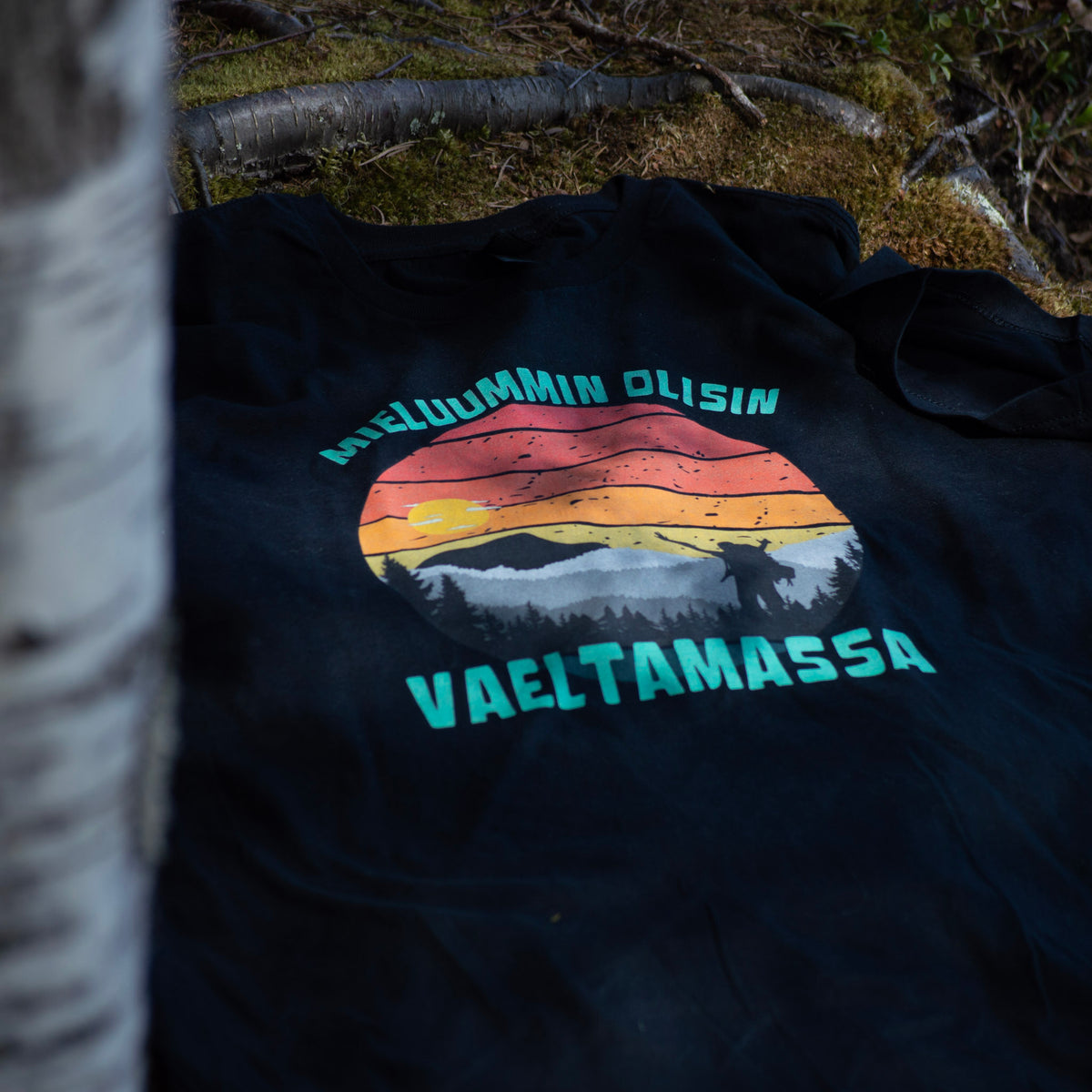 Musta Mieluummin olisin vaeltamassa -miesten t-paita tunturimaisemaprintillä, aseteltuna sammalelle.